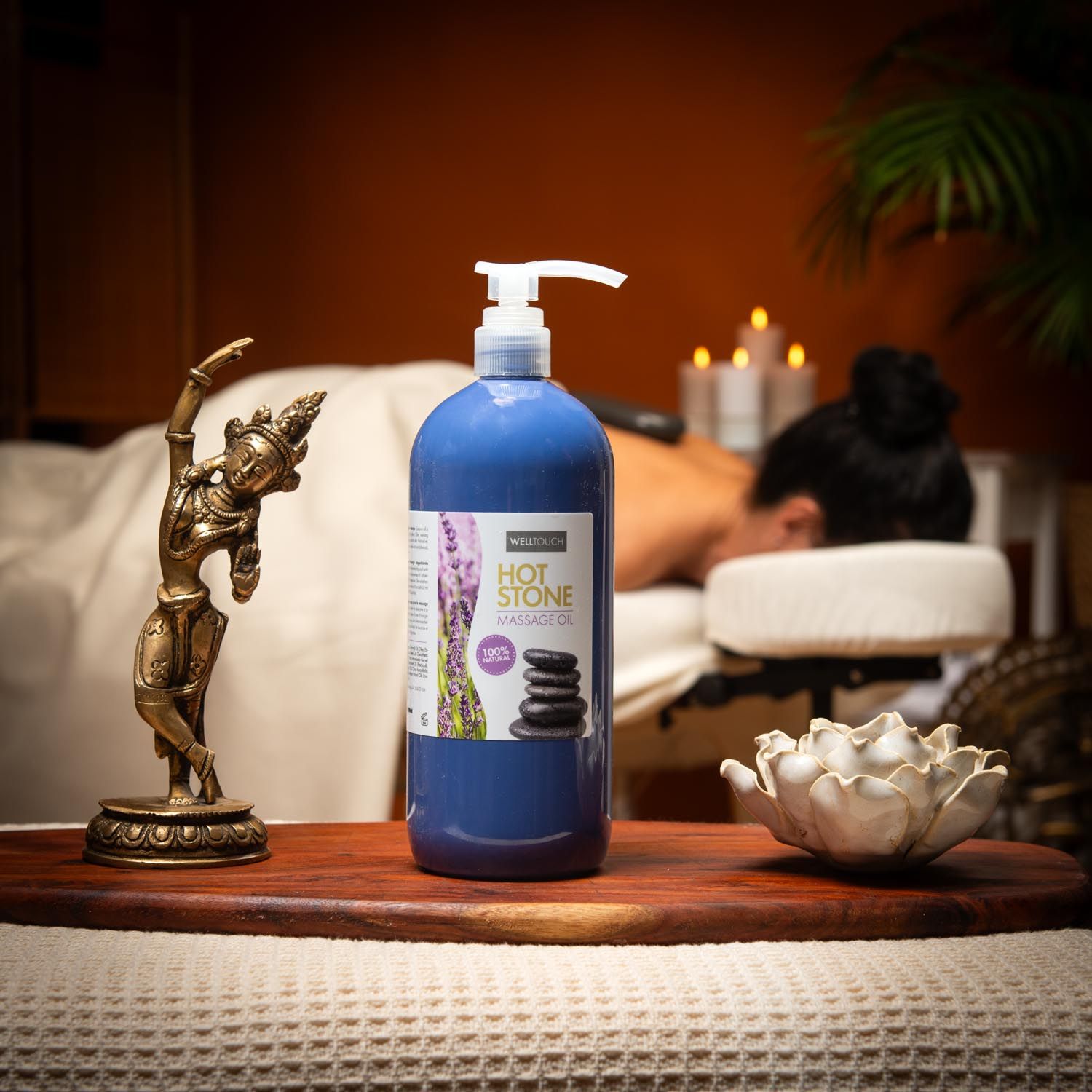 Blaue Flasche Welltouch Hot Stone Massageöl mit Spender. Auf Holztablett, neben Buddha-Figur und Lotusblüte. Im Hintergrund Person auf Massageliege.