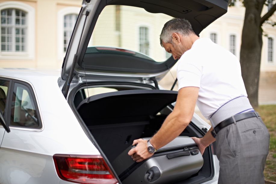 Mann lädt Koffer in Auto ein, trägt Bauerfeind LordoLoc Rückenorthese. Graues und blaues Design.