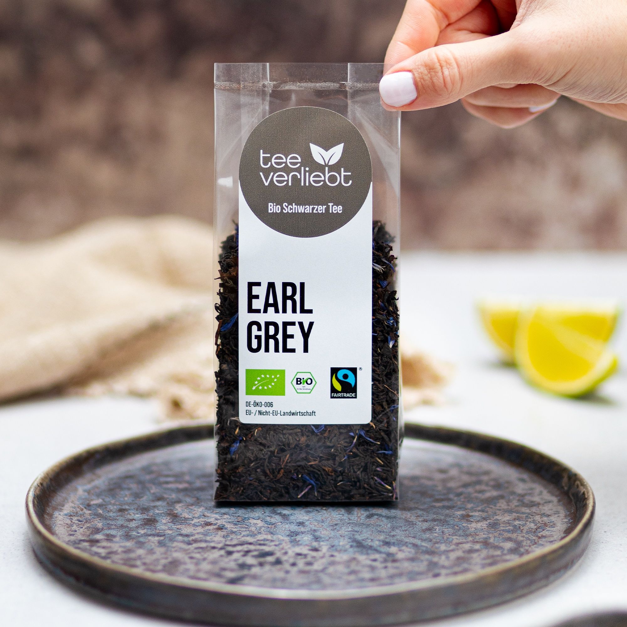 Hand hält transparente Verpackung mit losem Earl Grey Tee. Bio-Siegel und Fairtrade-Logo.