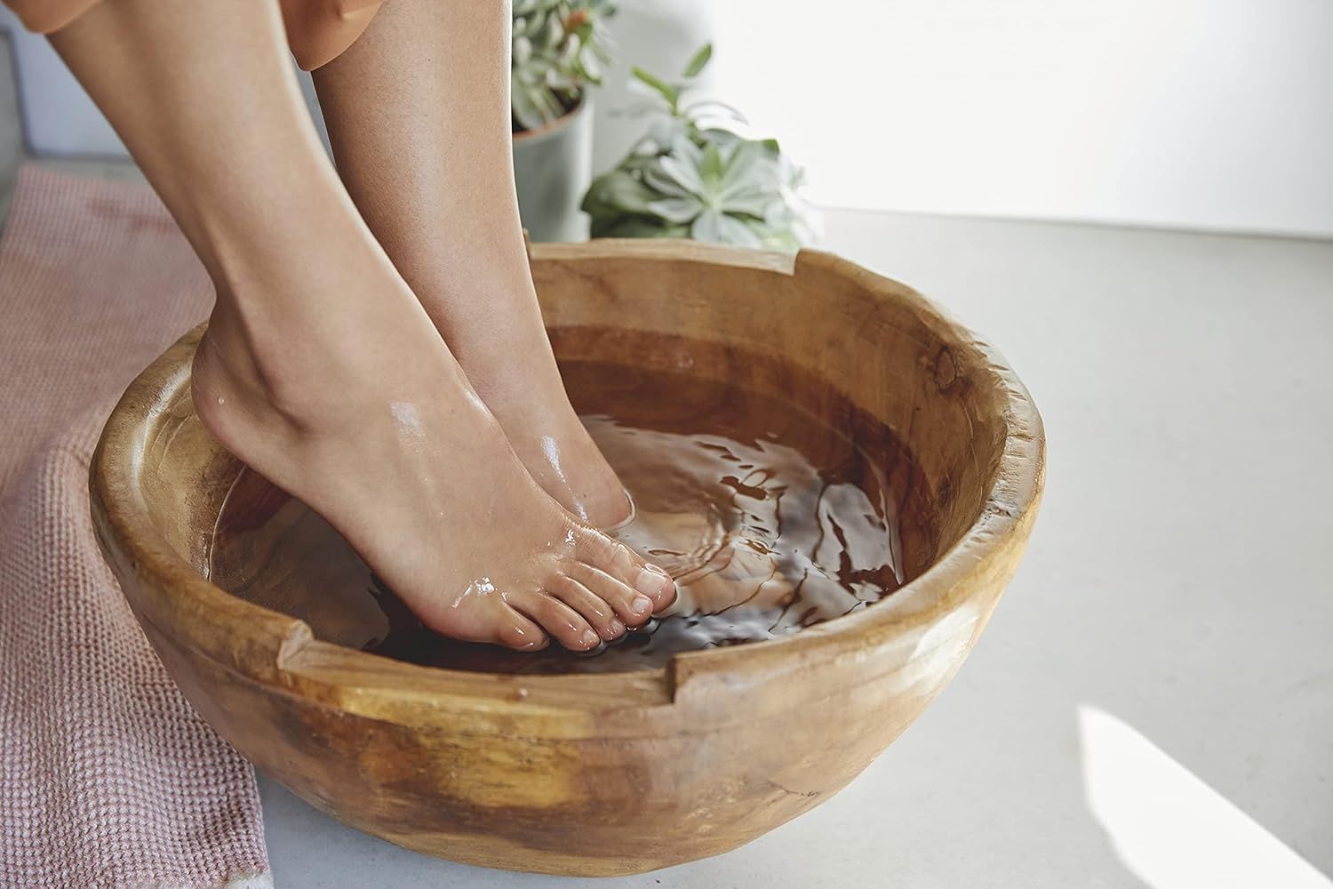 Füße in einer Holzschale mit Wasser. Helle Haut, leicht gerötete Füße. Hintergrund: Pflanzen, rosa Handtuch, heller Boden.