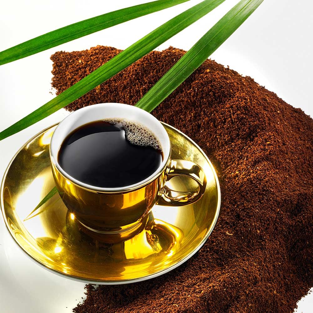 Goldene Tasse Kaffee auf Untertasse, umgeben von Kaffeepulver und grünen Blättern.