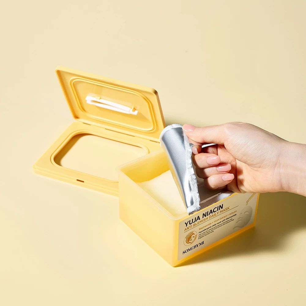 Geöffnete gelbe Maskenpackung. Hand entnimmt eine silberne Maskenverpackung. Aufschrift: Yuja Niacin Anti Blemish Daily Mask.