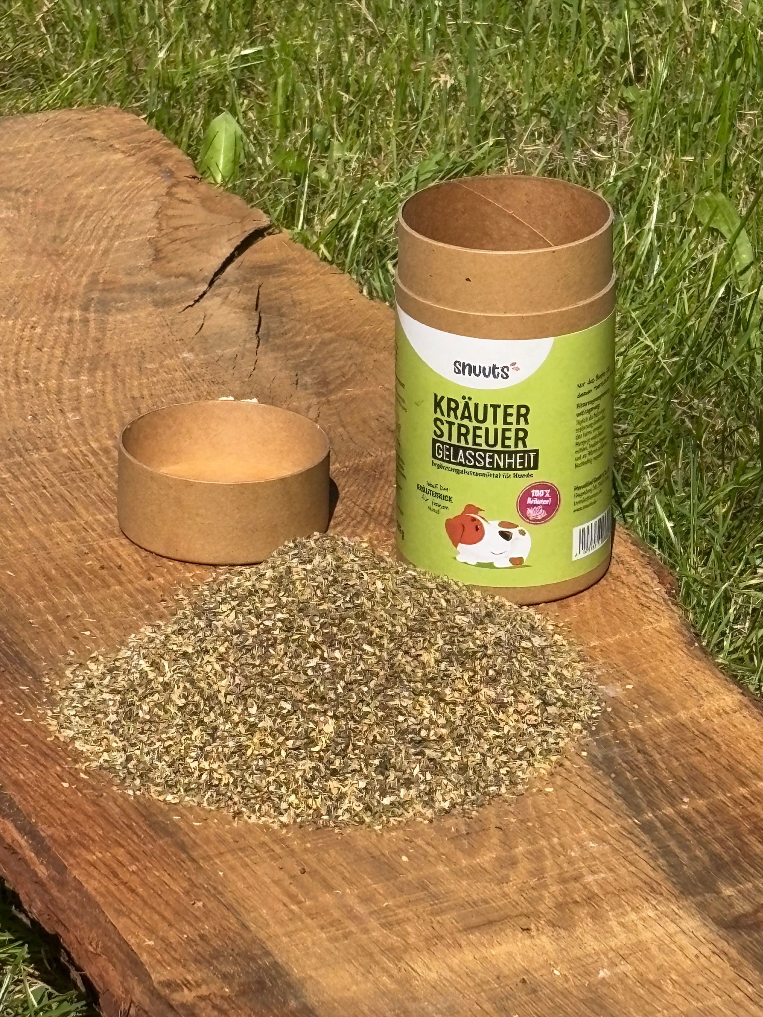 Kräuterstreuer-Dose, Deckel und Kräuterhaufen auf Holz. Dose mit grünem Etikett, Aufschrift: snuuts Kräuterstreuer Gelassenheit. Brauner Deckel und Kräuterhaufen.