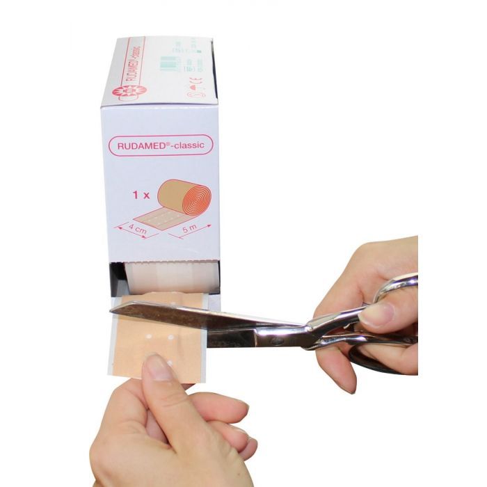 Rudamed-classic Wundpflaster-Verpackung. Schere schneidet Pflaster ab. Pflasterrolle sichtbar.