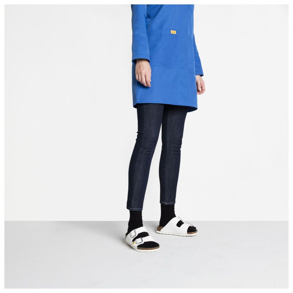 Person trägt weiße Sandalen. Blaue Jacke, dunkle Hose. Sandalen mit zwei Riemen, Marke Birkenstock.