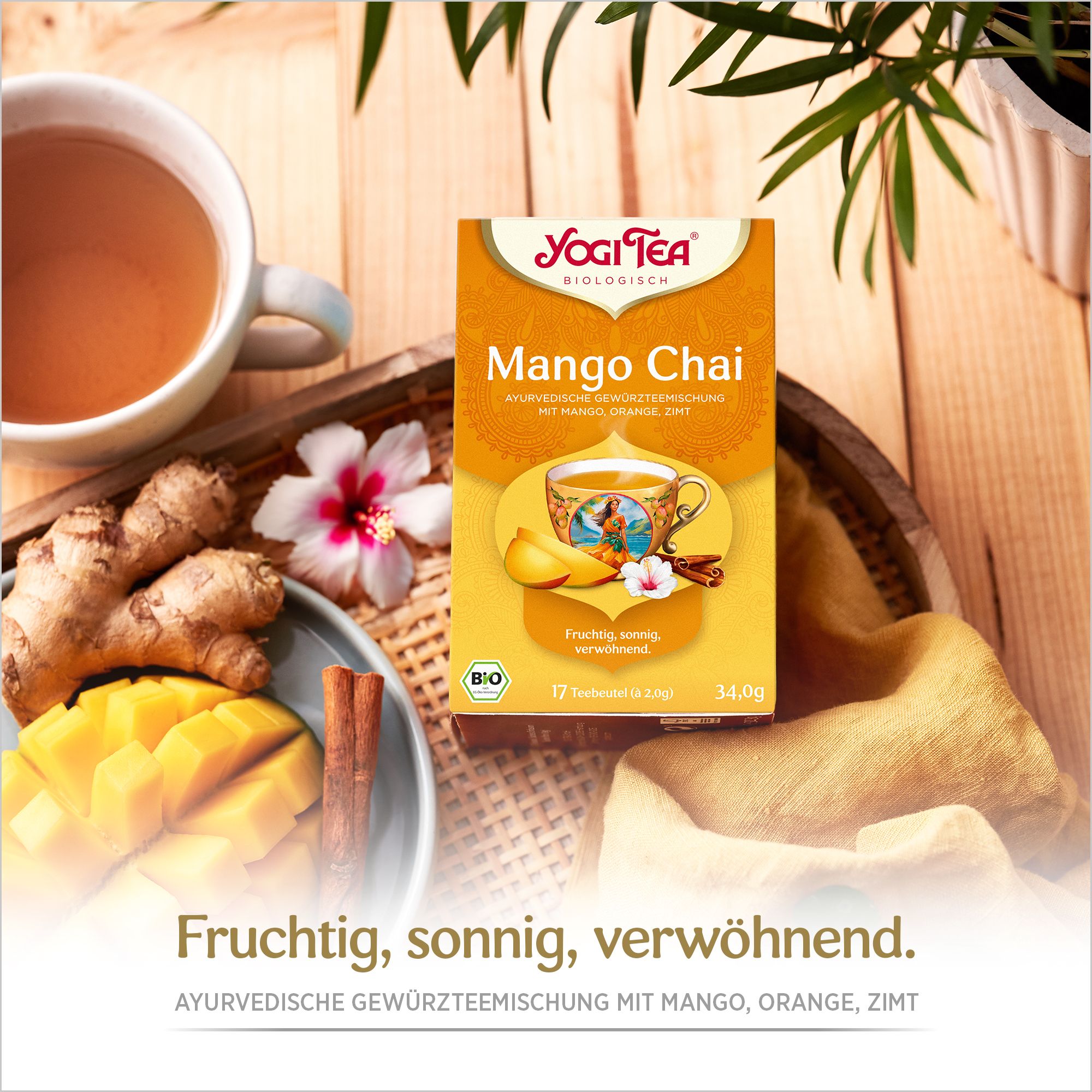 Mango Chai Tee-Verpackung auf einem Tablett. Neben der Packung: Tasse Tee, Mango, Ingwer, Zimtstange und Blüte. Text: Fruchtig, sonnig, verwöhnt.