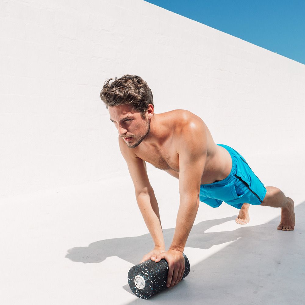 Mann stützt sich auf einer schwarzen Faszienrolle. Er macht Liegestütze. Blaue Shorts. Helle Umgebung.
