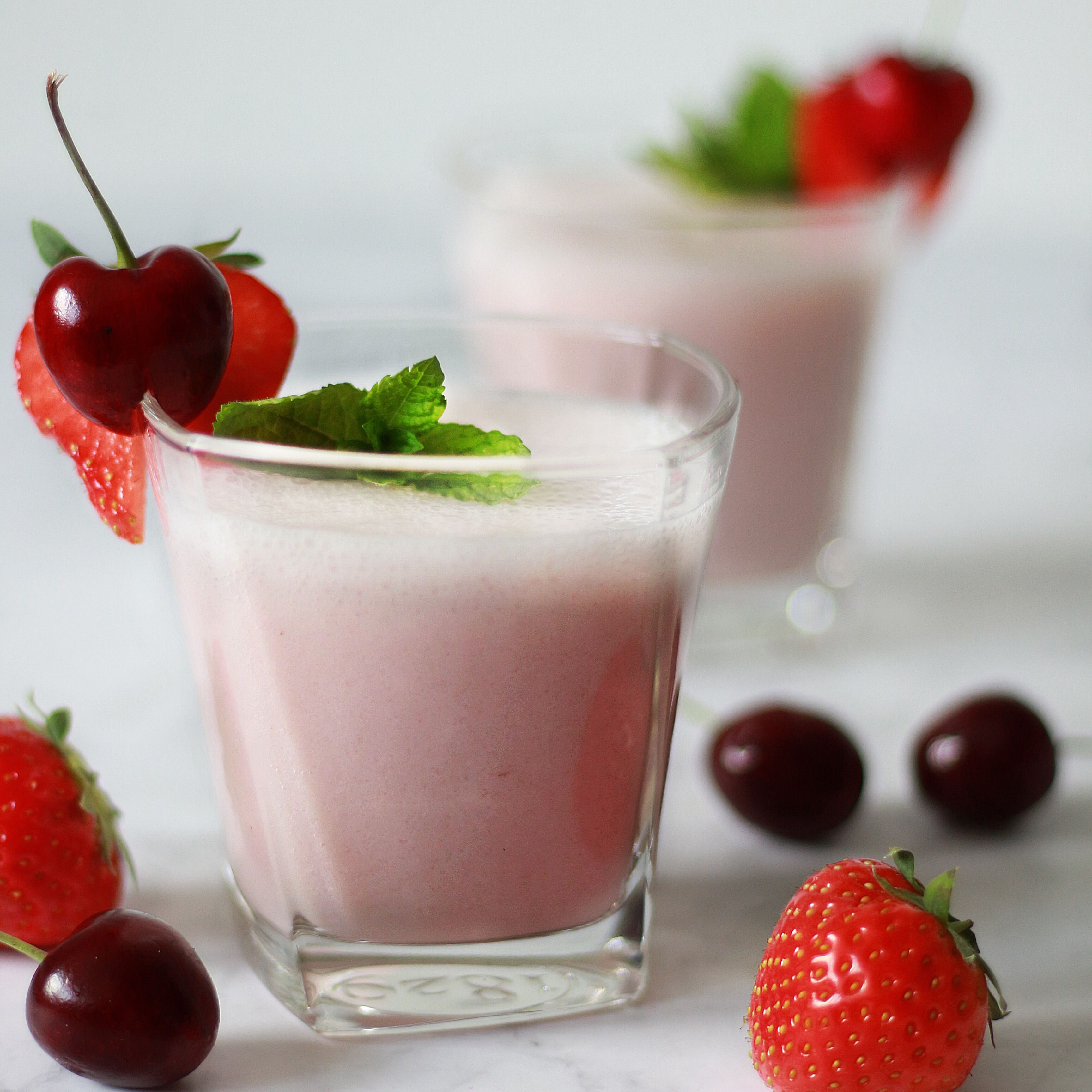 Zwei Gläser mit rosa Smoothie, Erdbeeren, Kirschen und Minze als Dekoration.