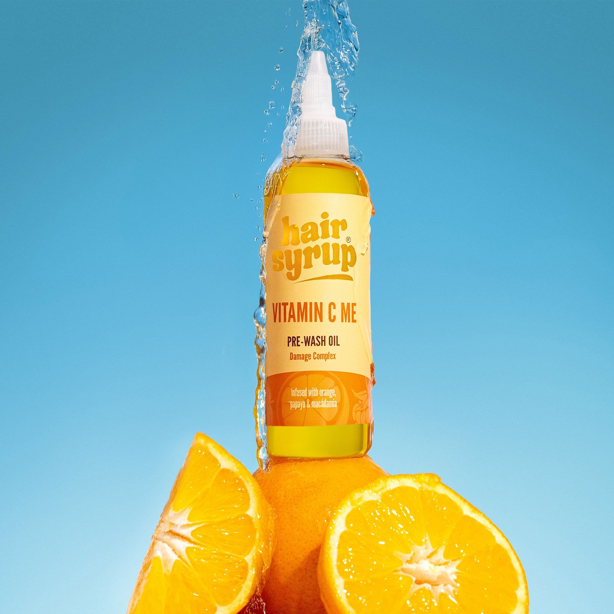 Gelbe Flasche mit "Hair Syrup Vitamin C Me"-Etikett. Orangen und Wasserstrahl im Hintergrund. Pre-Wash Oil.