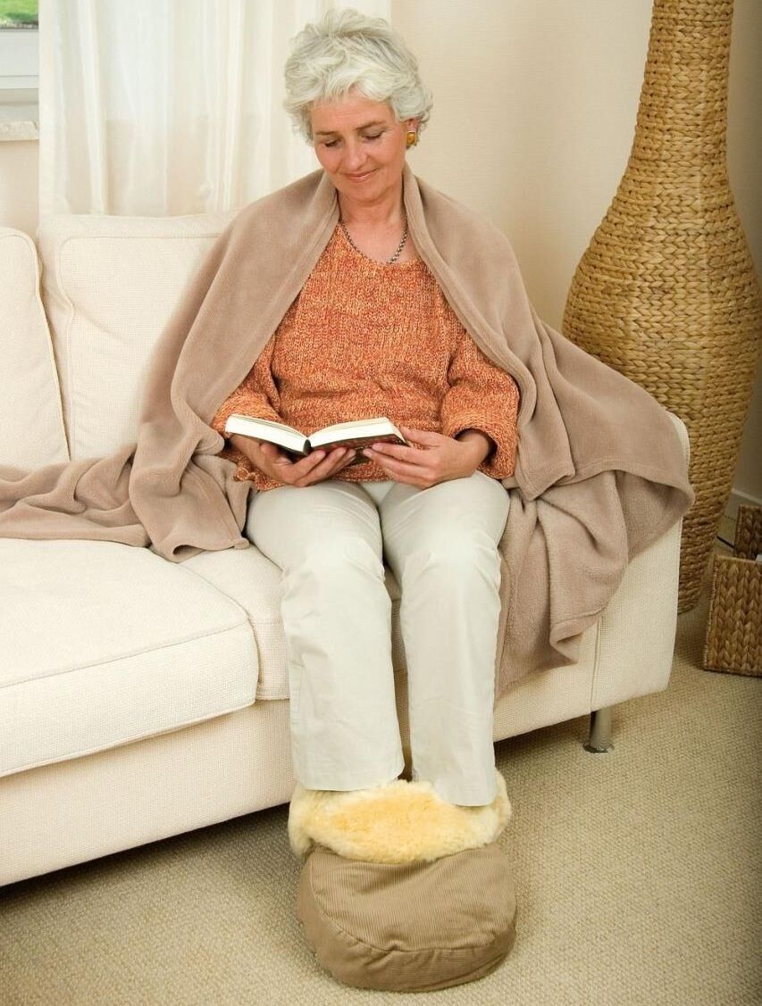 Ältere Frau sitzt auf einem Sofa, bedeckt mit einer Decke, Füße in einem Fußwärmer, hält ein Buch.