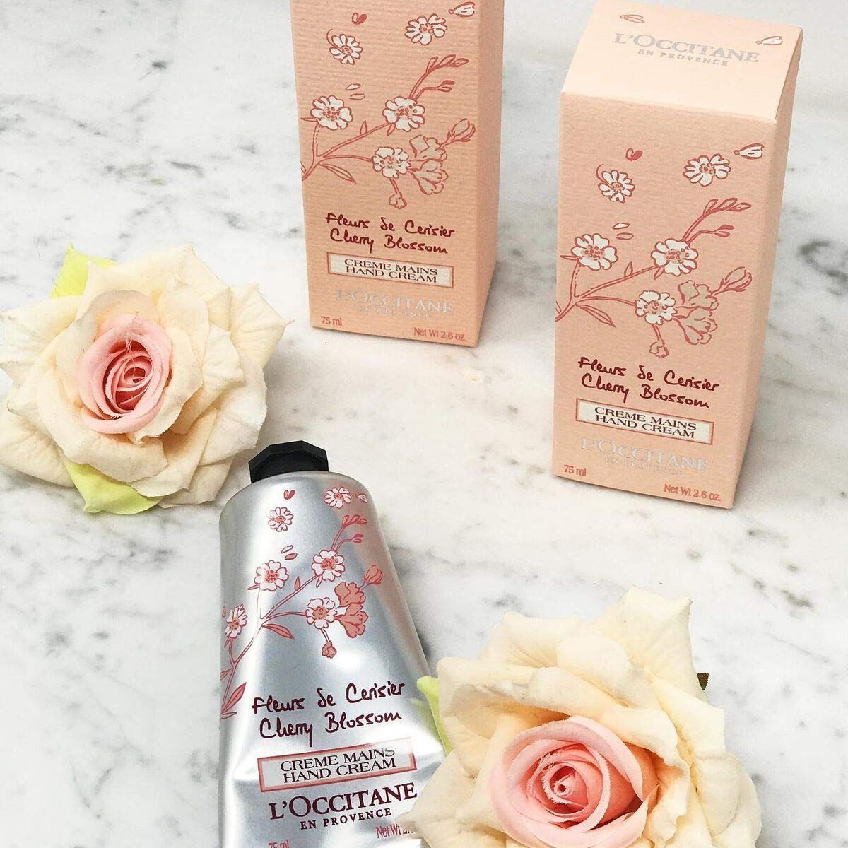 Produkt-Arrangement: Tube, zwei Schachteln, Rosen. Aufdruck: "Fleurs de Cerisier Cherry Blossom", "Crème Mains Hand Cream", "L'Occitane".