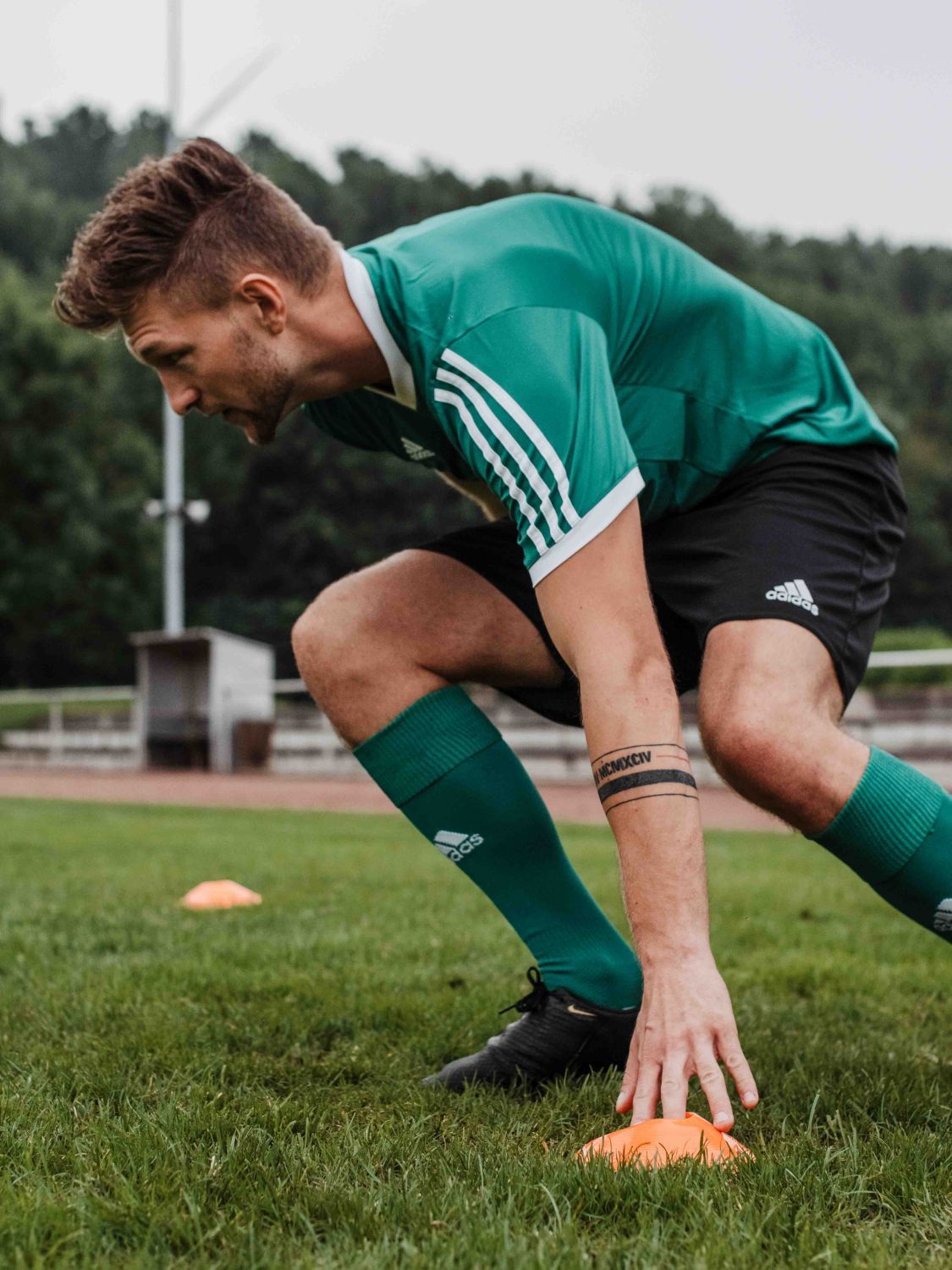Mann in grünem Trikot und schwarzen Shorts trainiert auf einem Rasen. Orangefarbene Hütchen im Vordergrund.