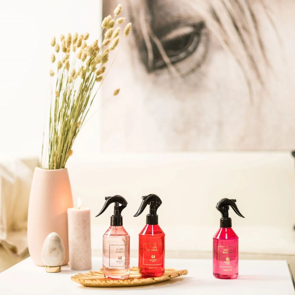 Drei Sprühflaschen auf Tablett. Schwarze Sprühköpfe, rosa Flüssigkeit. Etiketten mit Text. Dekoration: Vase, Kerze.