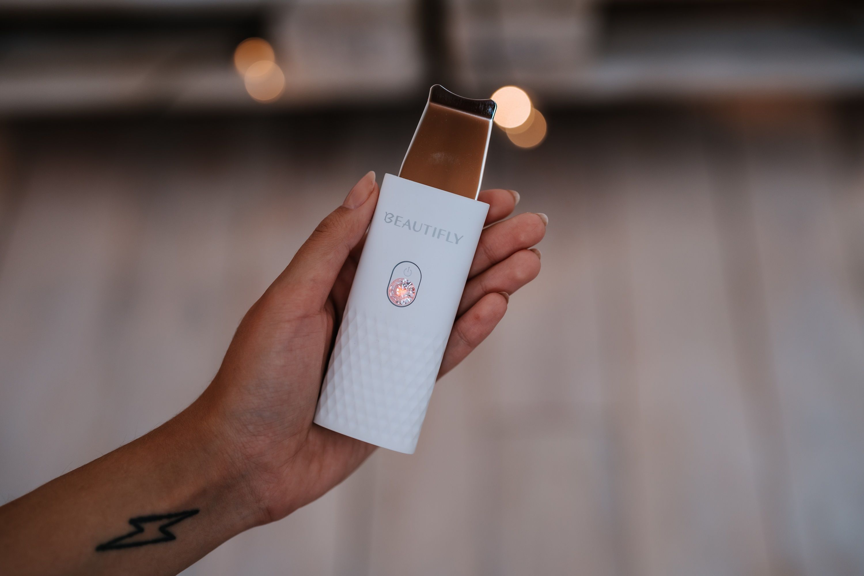 Hand hält BEAUTIFLY B-Scrub Mini. Weißes Gerät, silberne Spitze, rosa Knopf. Geometrisches Muster.