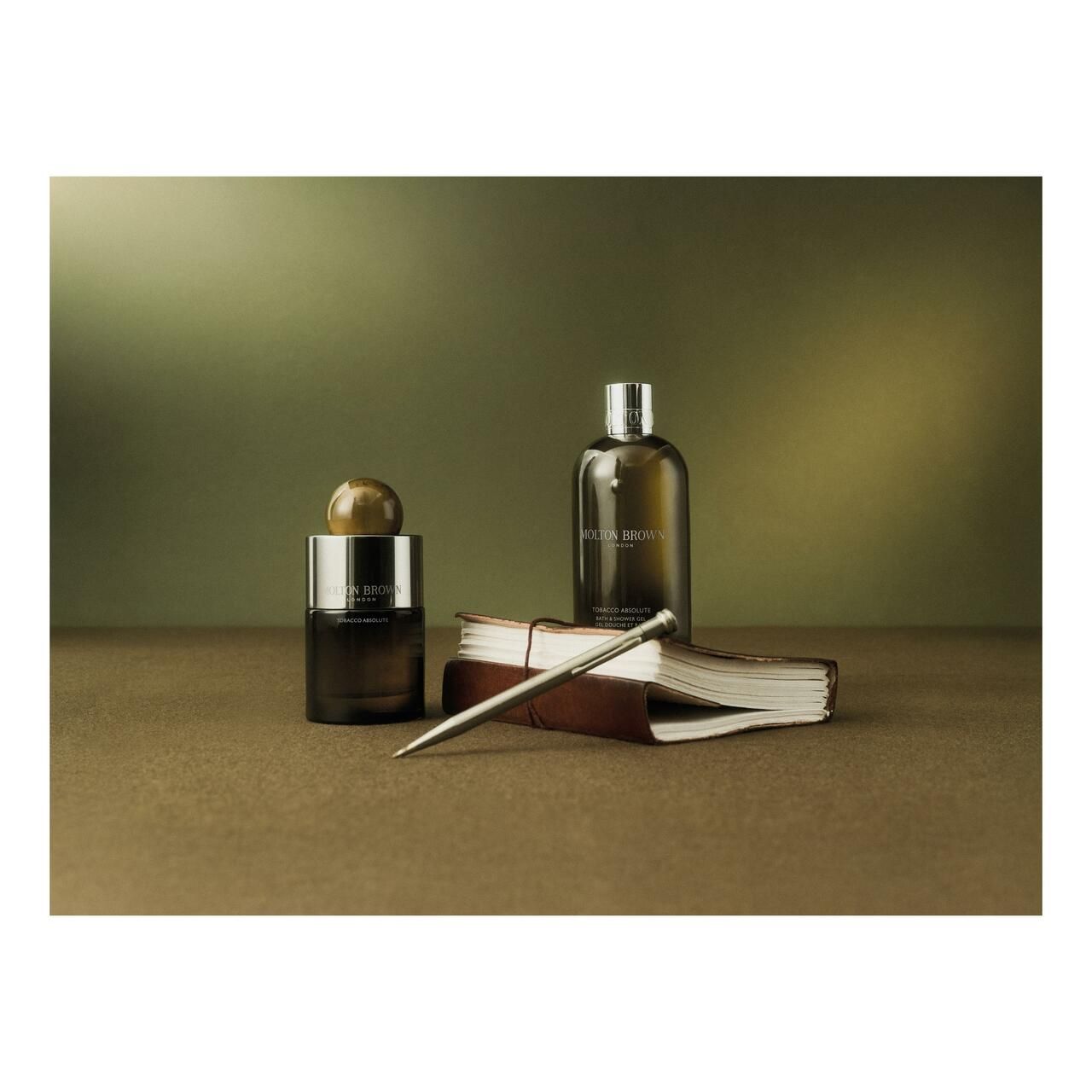Zwei Flaschen, ein Buch, ein Stift. Aufschrift: Molton Brown. Hintergrund: Grün und Braun.