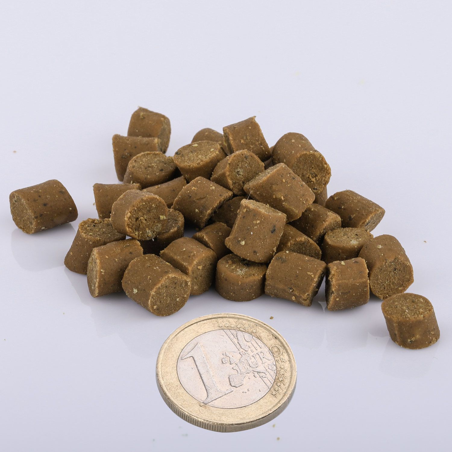 Haufen brauner, würfelförmiger Snacks. Daneben eine 1-Euro-Münze. Helle, neutrale Oberfläche.