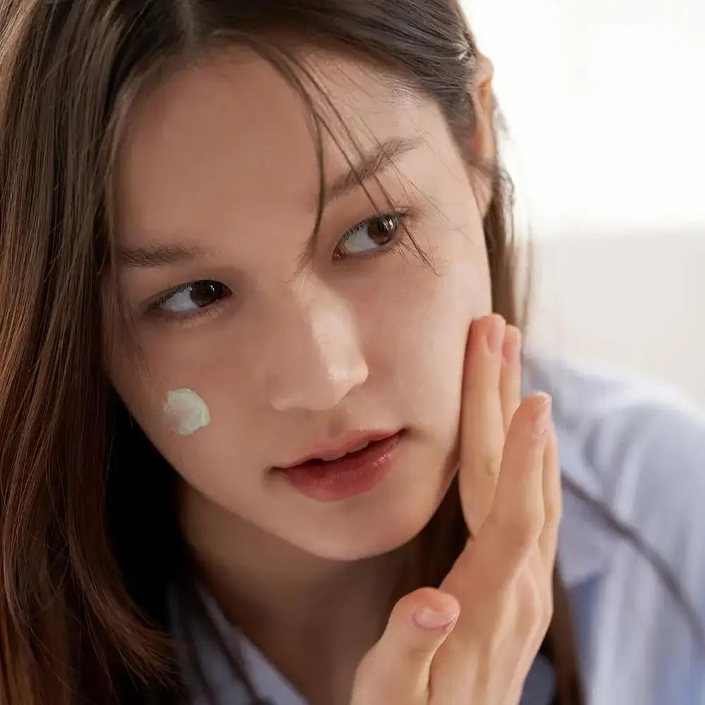 Frau trägt grüne Make-up-Grundierung auf die Wange auf. Helle Haut, braune Augen, blaue Bluse.