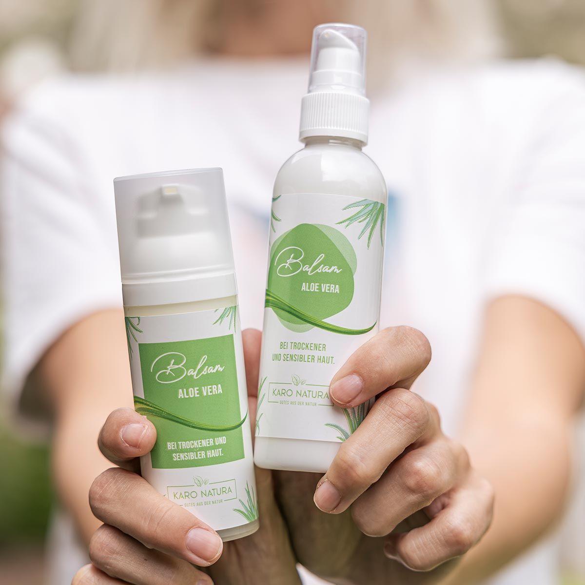 Zwei weiße Spender mit grünem Etikett. Aufschrift: Balsam Aloe Vera. Text: Bei trockener und sensibler Haut. Marke: Karo Natura. Gehalten von Händen.