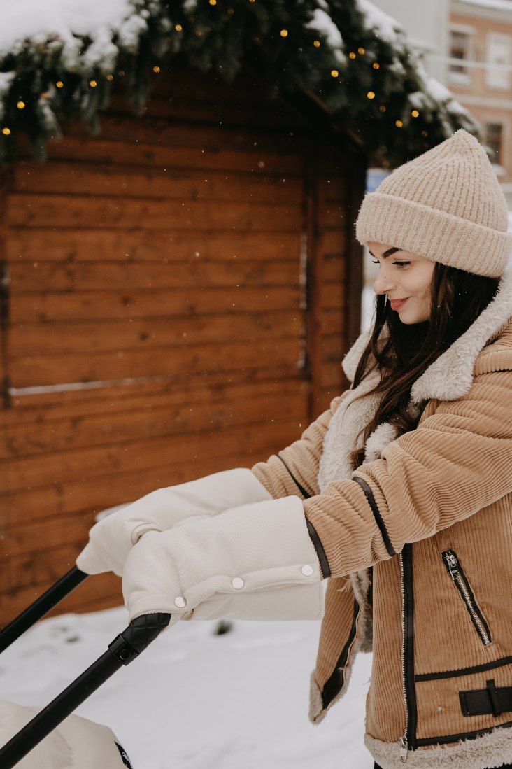 Frau schiebt Kinderwagen im Schnee, trägt beige Mütze und Cordjacke.