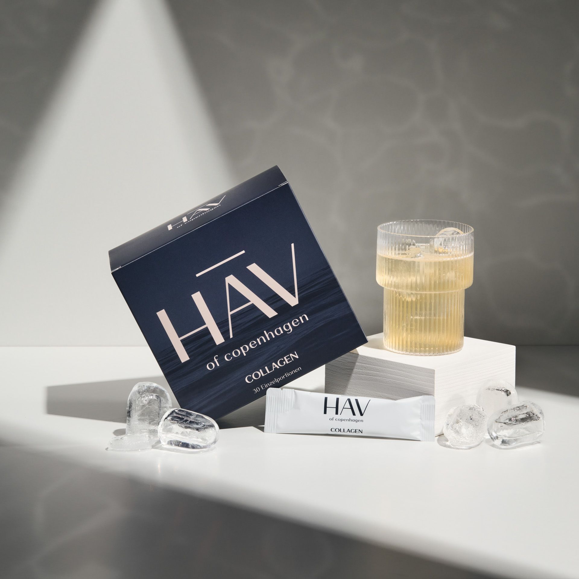 Produktverpackung, Glas mit Eiswürfeln und Einzelportion HAV Collagen-Stick auf weißem Podest.