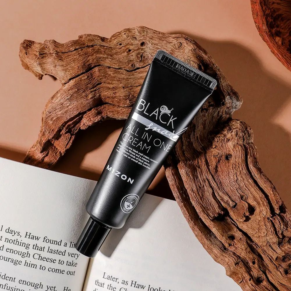 Schwarze Tube mit Aufschrift BLACK snail ALL IN ONE CREAM und MIZON. Auf Holz und Buch.