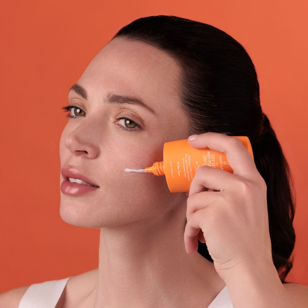 Frau trägt Creme aus orangefarbener Tube auf ihr Gesicht auf. Text: YOUTH LAB, City Guard Anti-Pollution Day Cream SPF50.