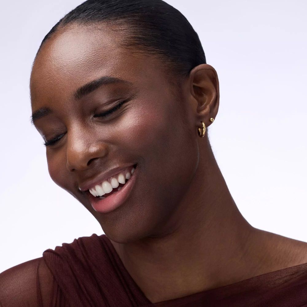 Porträt einer lächelnden Frau mit rosa Rouge auf den Wangen. Sie trägt goldene Ohrringe.