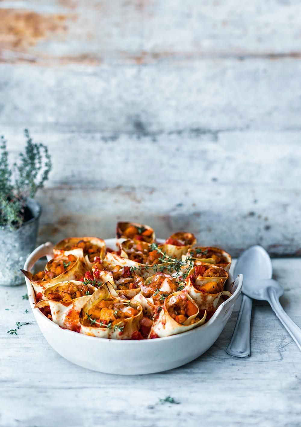 Weiße Schüssel mit Gemüse-Rollen. Tomaten, Gemüse, Kräuter. Auf hellem Holz. Löffel. Topf mit Kräutern.