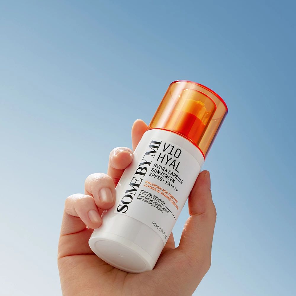 Weißer Flakon mit orangefarbenem Deckel. Aufschrift: SOME BY MI V10 Hyal Hydra Capsule Sunscreen SPF50+ PA++++. Hand hält Flakon vor blauem Himmel.