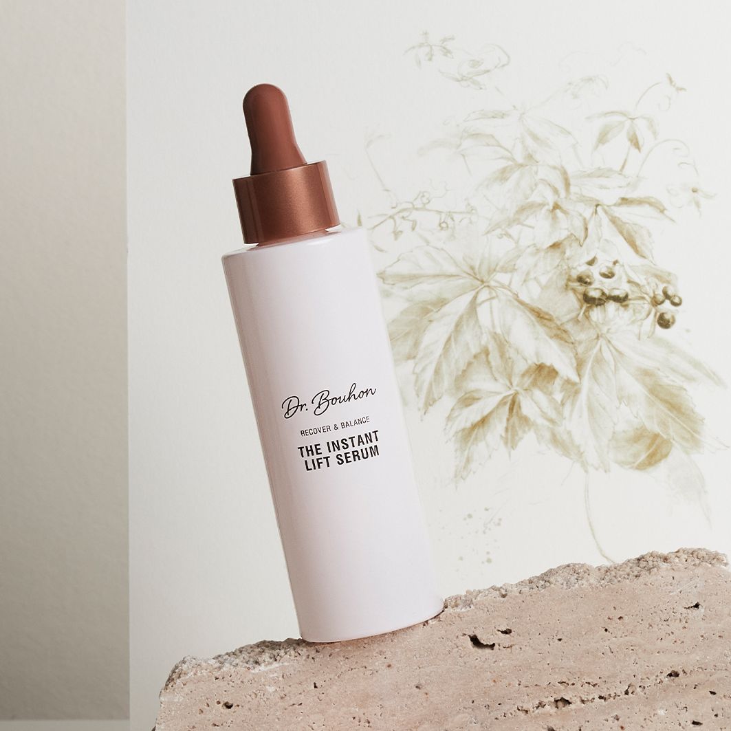 Flacon rose avec bouchon marron sur pierre. Inscription: Dr. Bouhon, THE INSTANT LIFT SERUM. Fond: plante.