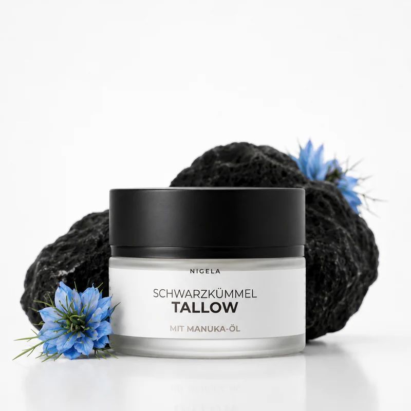 Glas mit schwarzem Deckel, Etikett 'Nigella Schwarzkümmel Tallow mit Manuka-Öl', auf dunklem Gestein mit blauen Blüten.