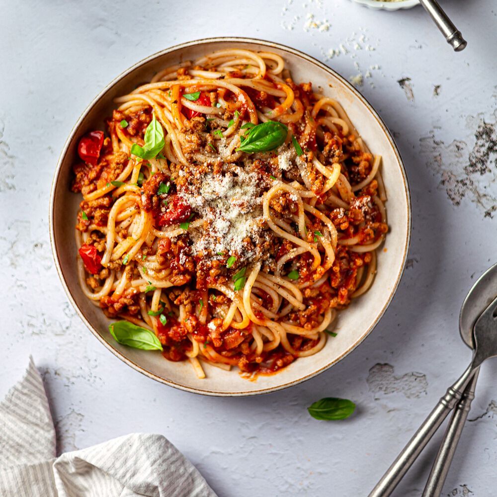 Teller mit Spaghetti mit Tomaten-Fleisch-Sauce, Basilikum und Käse. Besteck liegt daneben. Auf einem Tuch.