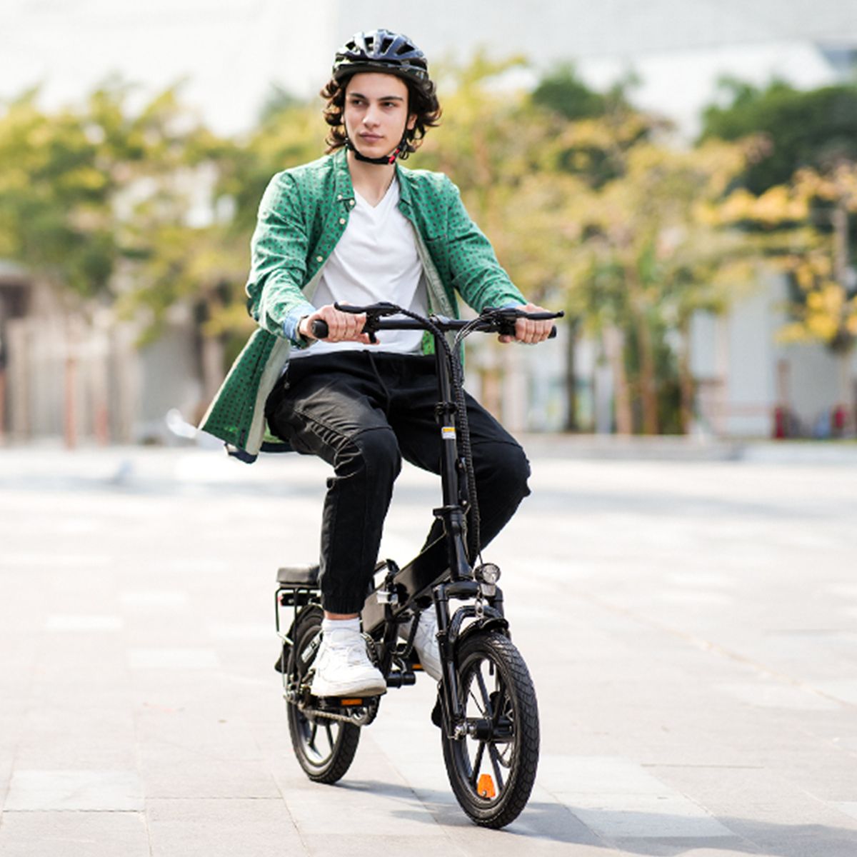 Person fährt auf einem schwarzen, zusammenklappbaren Elektrofahrrad. Trägt Helm und grüne Jacke. Hintergrund: Bäume.