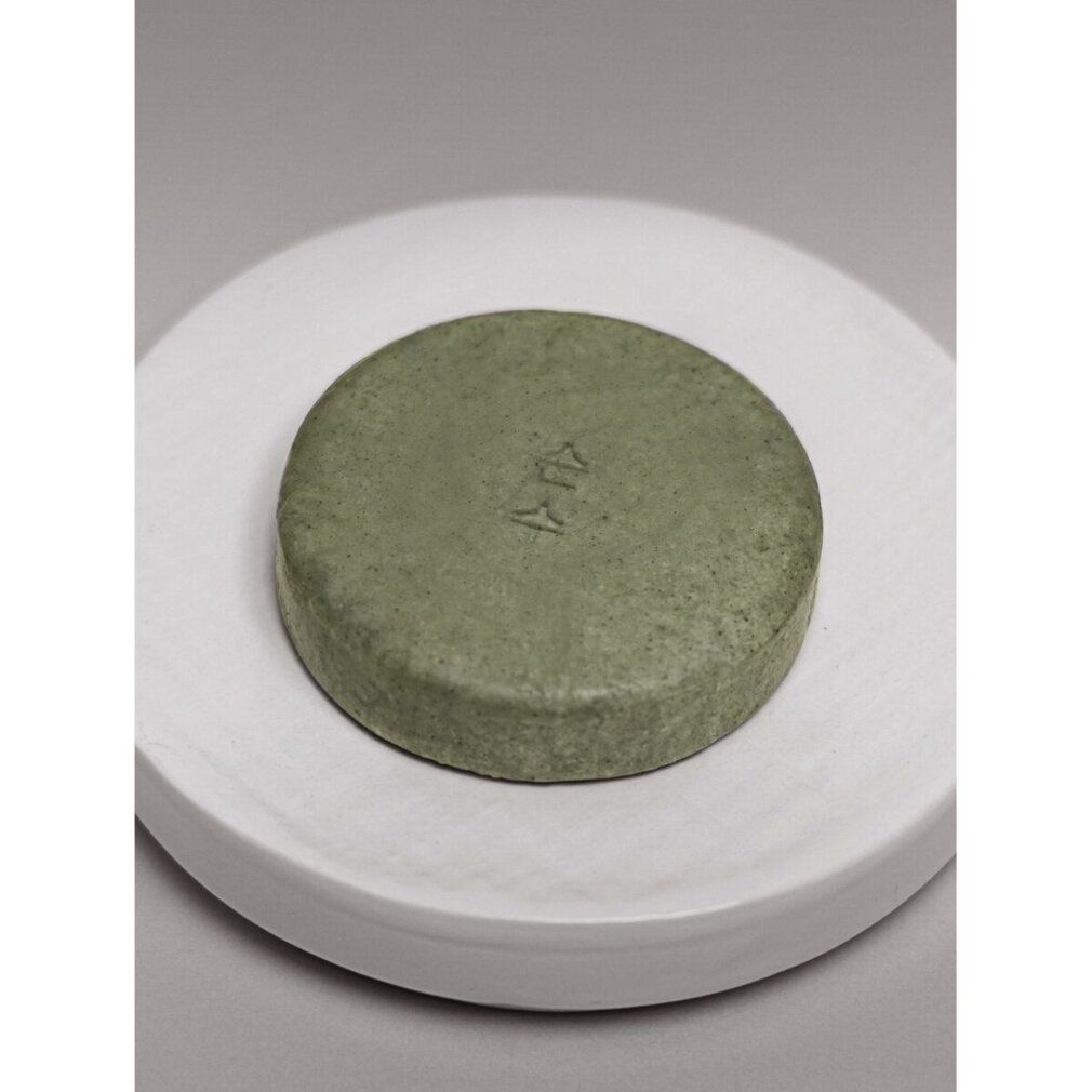 Runder, grüner Gegenstand auf weißer, runder Unterlage. Oberfläche strukturiert. Auf grauem Hintergrund.
