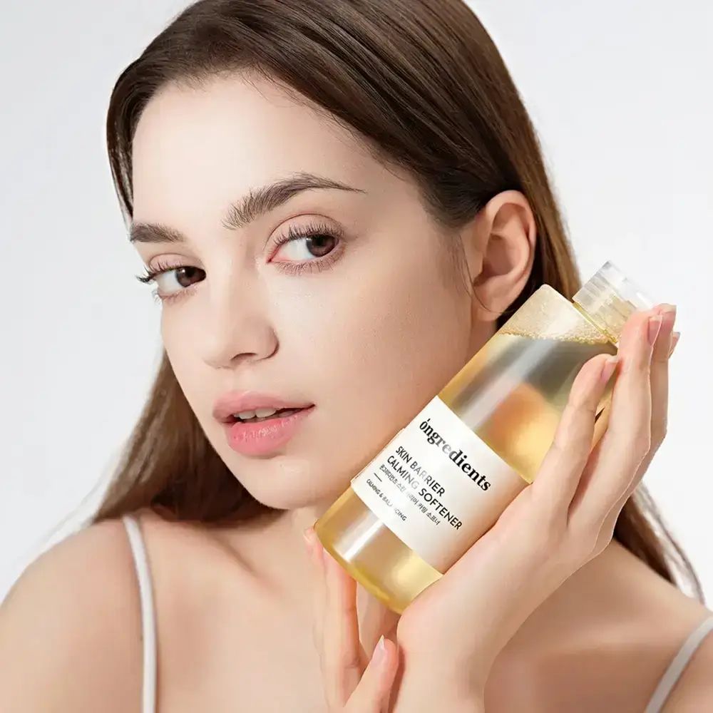Eine Frau hält eine transparente Flasche mit gelblicher Flüssigkeit. Auf der Flasche steht "ongredients SKIN BARRIER CALMING SOFTENER".