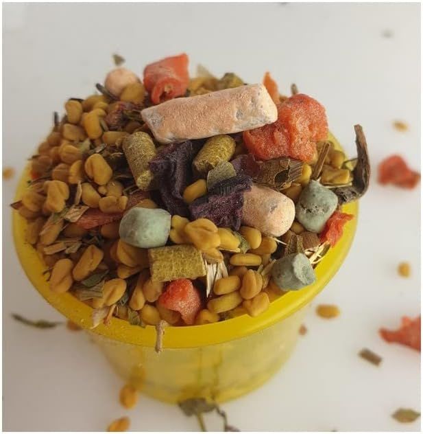 Gelber Becher gefüllt mit verschiedenen Futterbestandteilen. Enthält Samen, Gemüse und andere Zutaten.