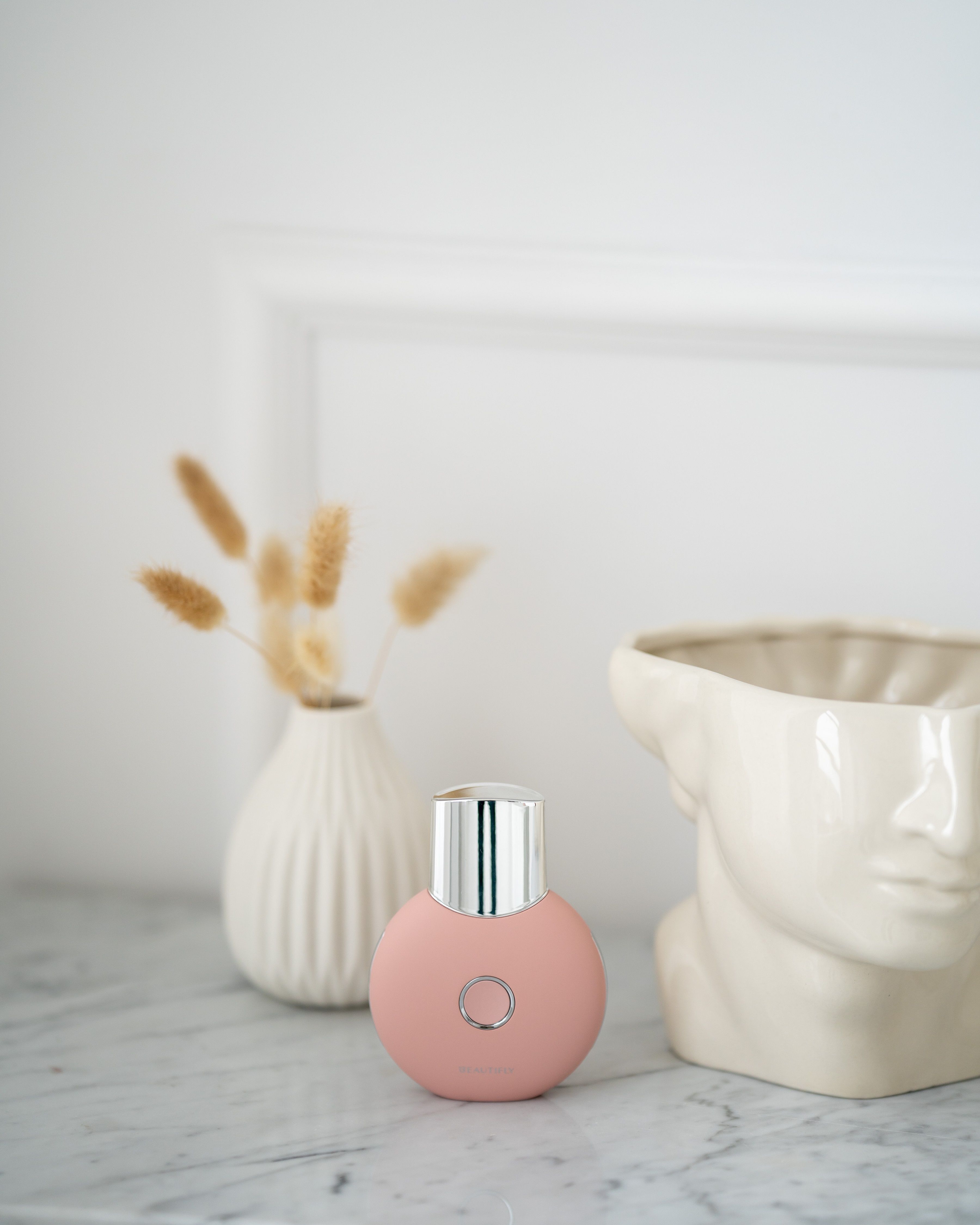 Rosa Hautpeeling-Gerät mit Metallaufsatz, platziert auf einer Marmoroberfläche. Neben einer Vase und einer Büste.