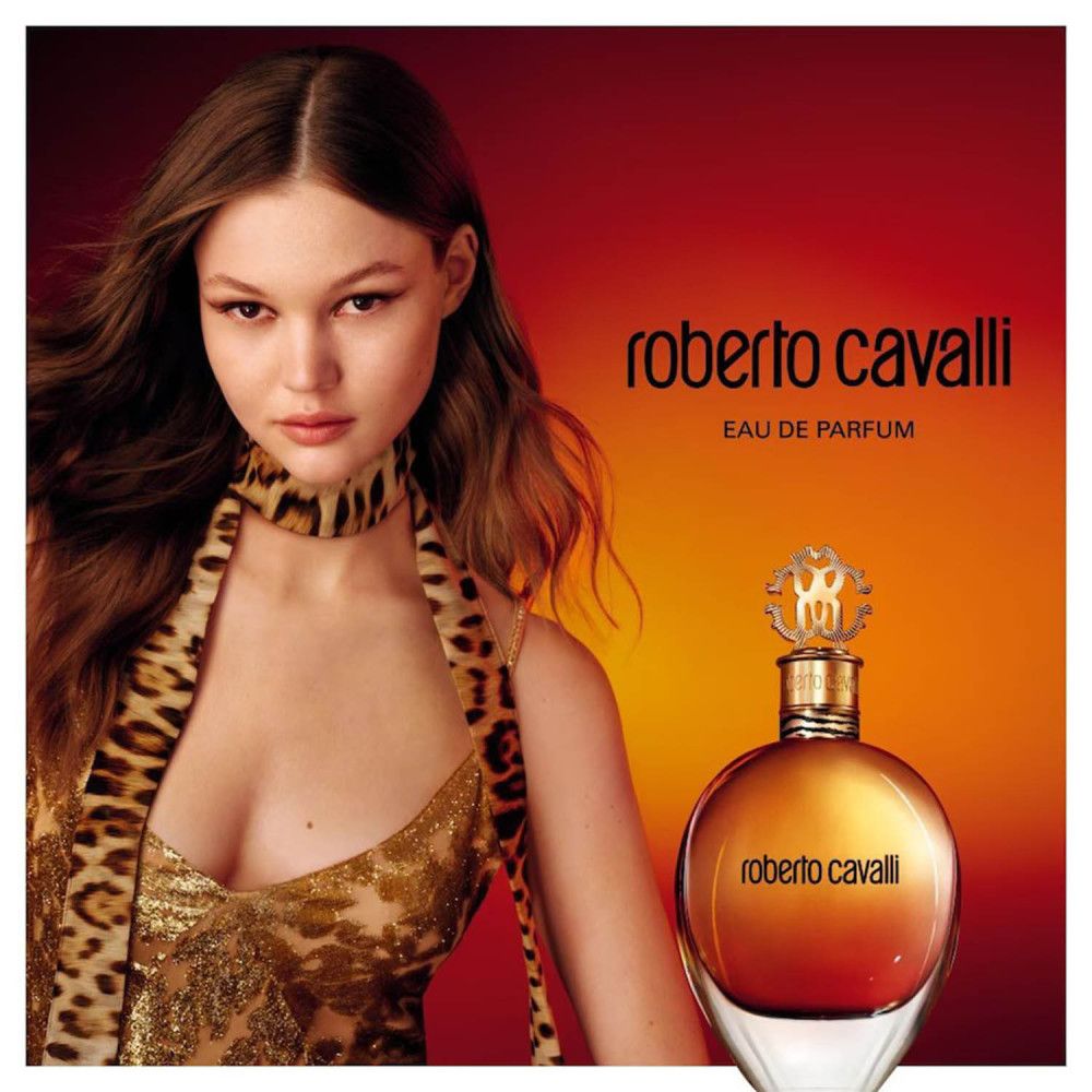 Werbefoto mit Frau und Flakon. Der Flakon ist rund, mit goldenem Verschluss und Schriftzug. Hintergrund rot-orange.