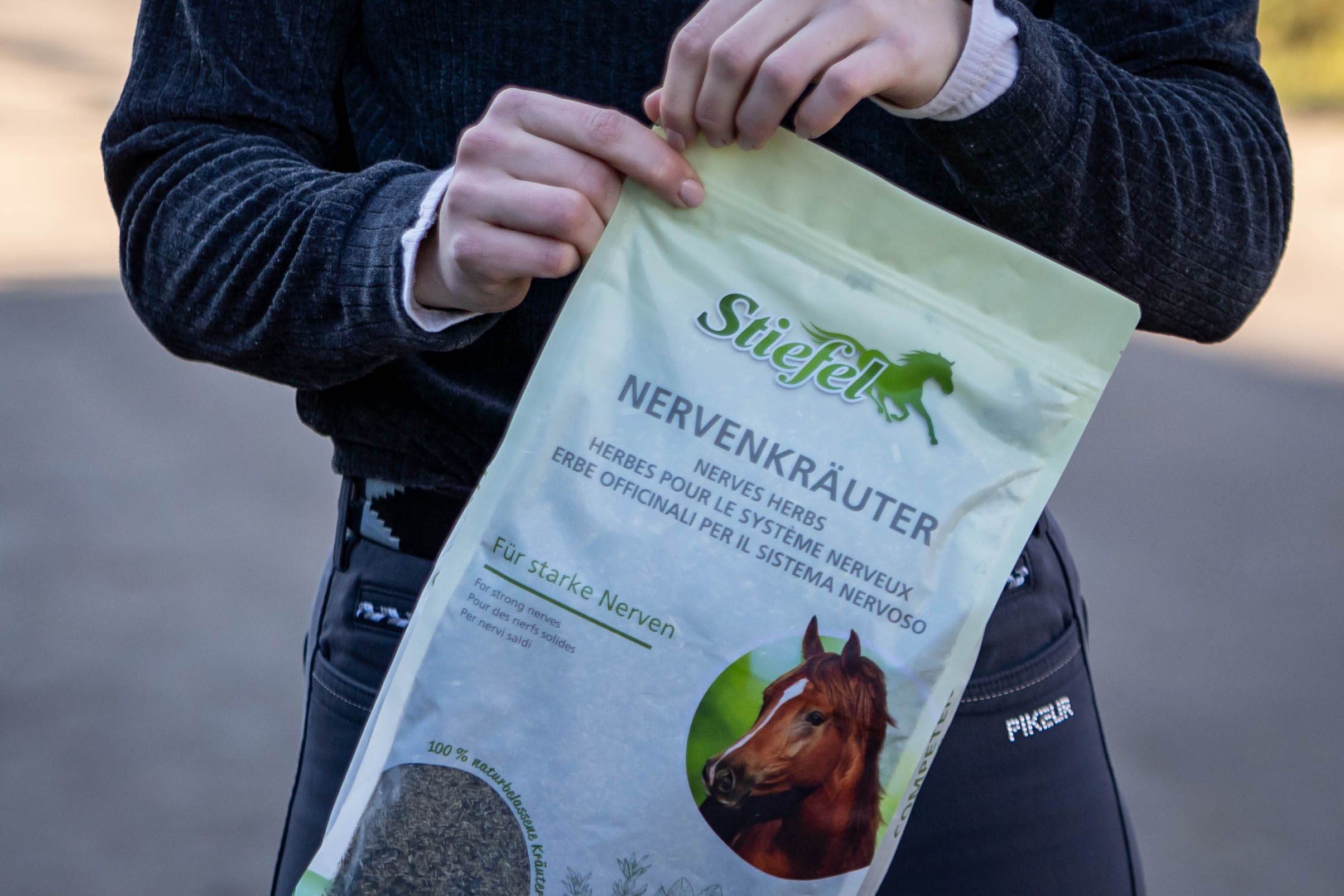 Person hält eine grüne Verpackung von Stiefel Nervenkräuter. Aufdruck: Produktname, Pferdeabbildung, Text in Deutsch, Französisch, Italienisch. Hintergrund: unscharf.