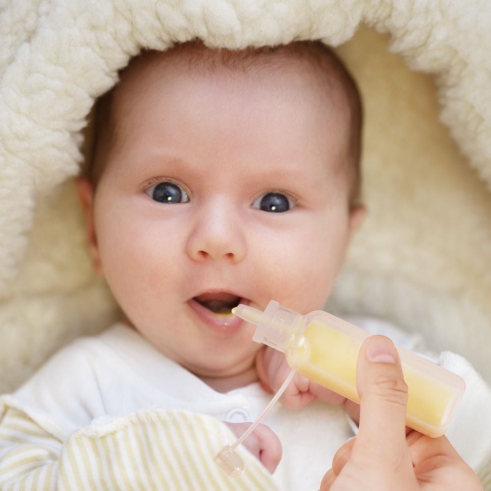Baby erhält Flüssigkeit aus einem transparenten Behälter. Der Behälter wird von einer Hand gehalten.