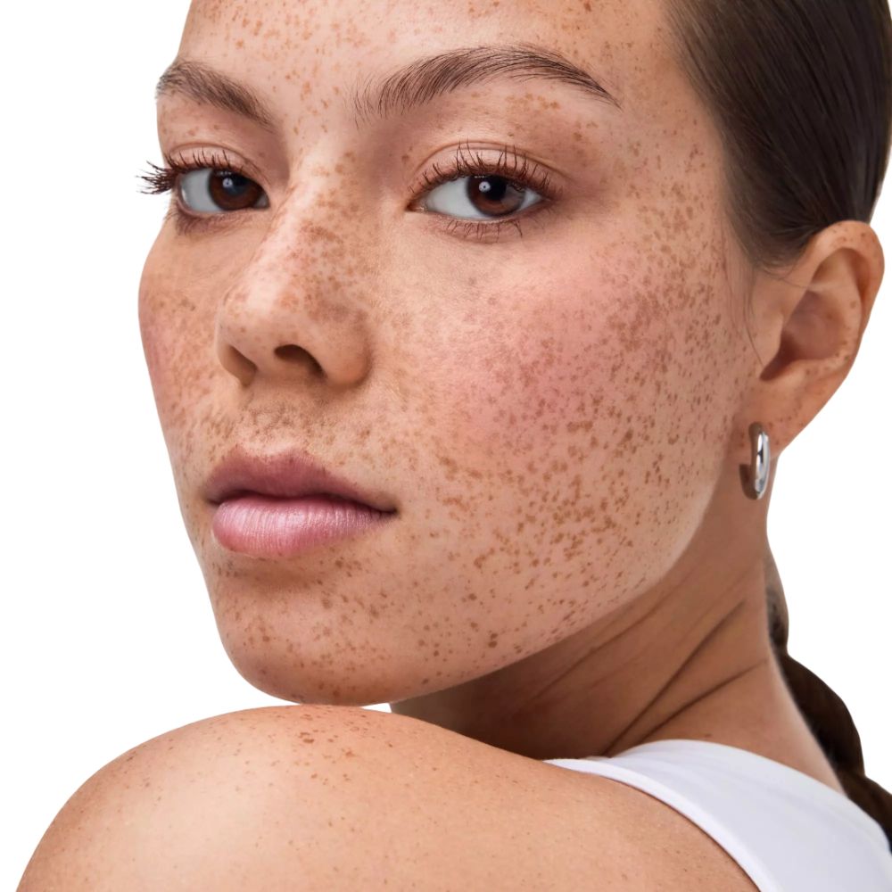 Nahaufnahme einer Frau mit Sommersprossen. Sie trägt einen weißen Tanktop und Ohrringe. Blick zur Kamera.