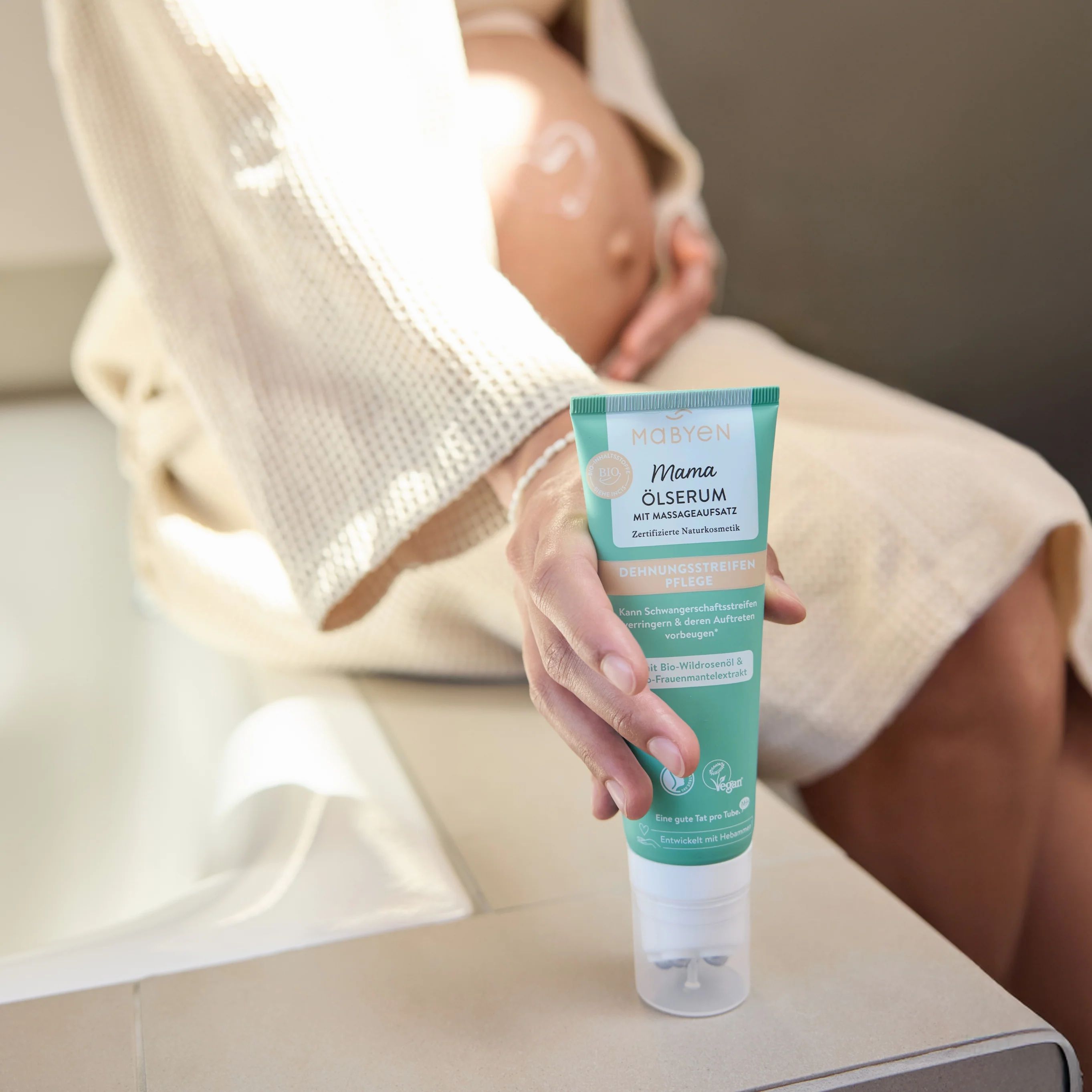 Schwangere Frau sitzt. Hält Tube. Creme auf Bauch. Türkis-weiße Tube.