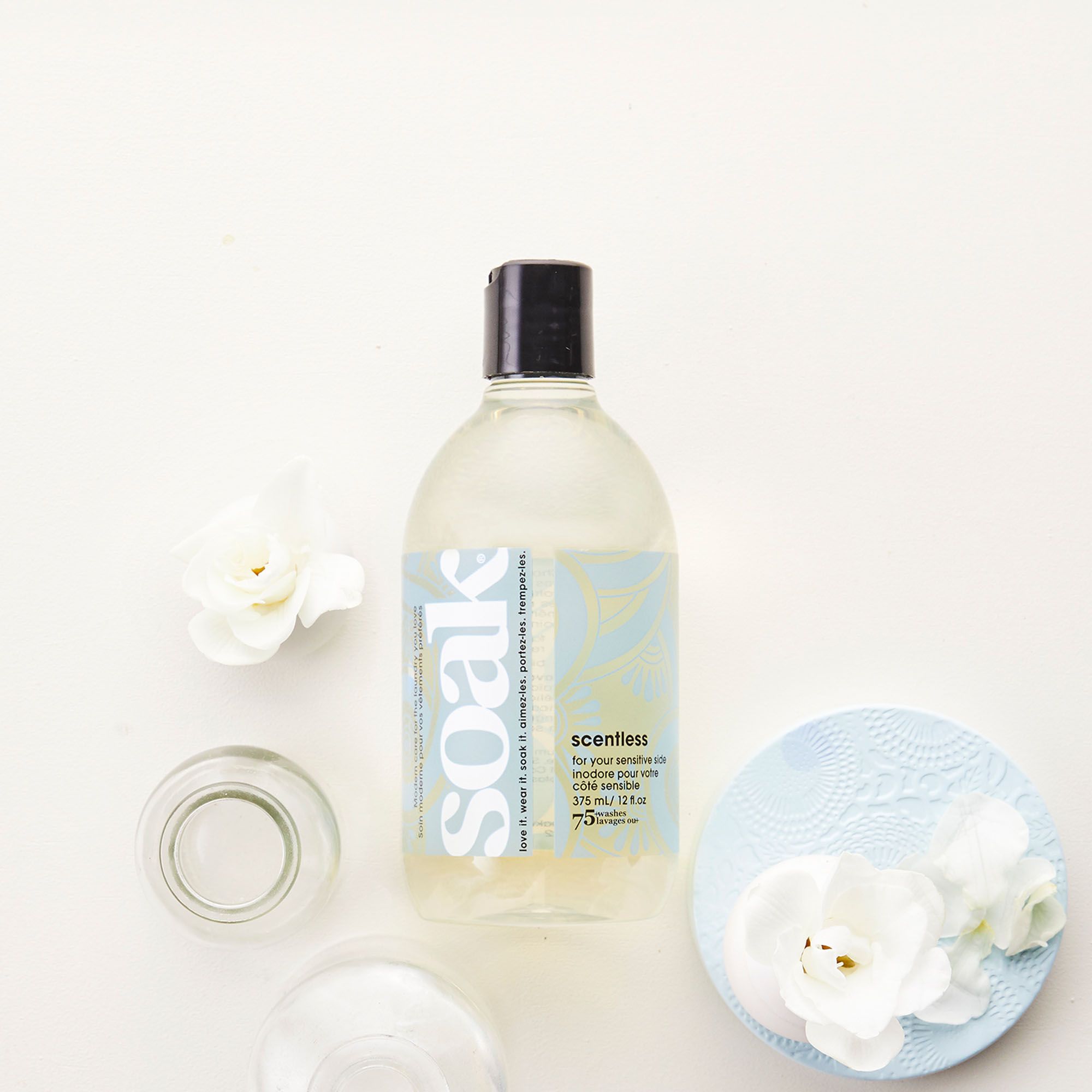 Soak Waschmittel, transparente Flasche mit schwarzem Deckel. Aufschrift: scentless. Neben Blumen und einem blauen Untersetzer.