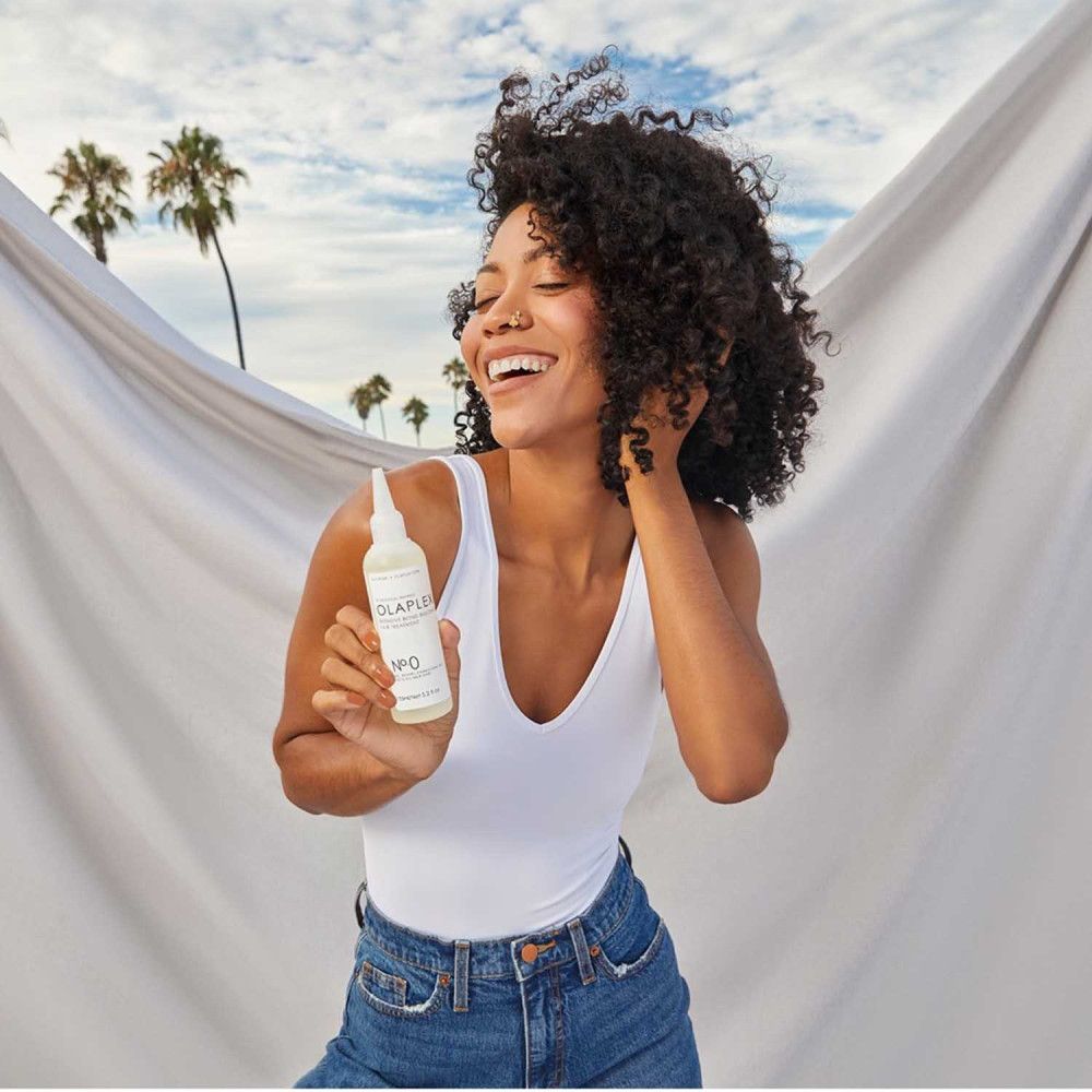 Frau hält eine Flasche OLAPLEX No. 0. Aufschrift: OLAPLEX Intensive Bond Building Hair Treatment. Hintergrund: Strand.