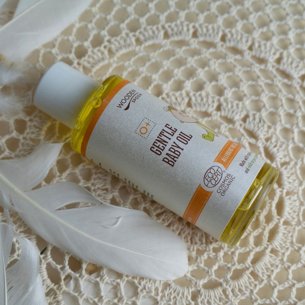 Babyöl-Flasche auf cremefarbenem Hintergrund mit Federn. Aufschrift: Gentle Baby Oil, Wooden Spoon, Cosmos Organic.