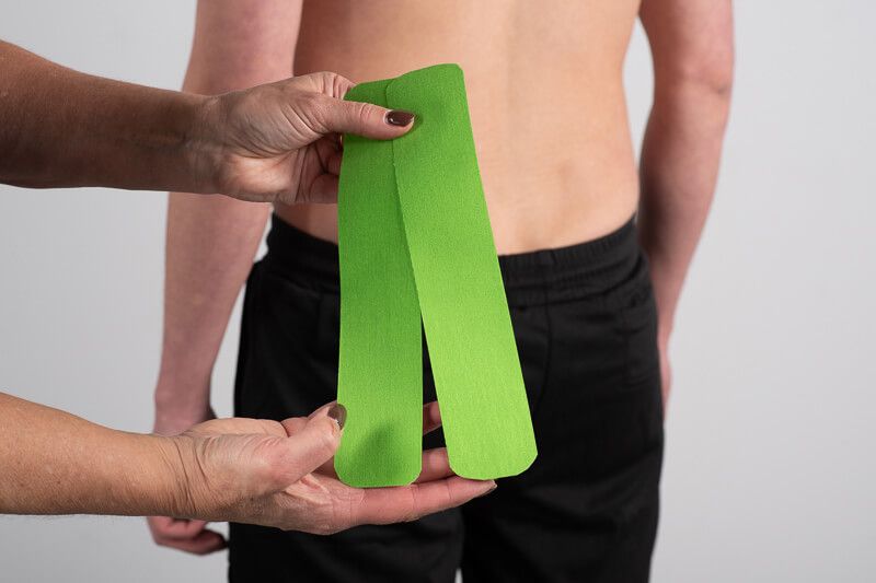Hände halten zwei grüne Streifen PhysioTape No.1. Person im Hintergrund. Helle Haut.