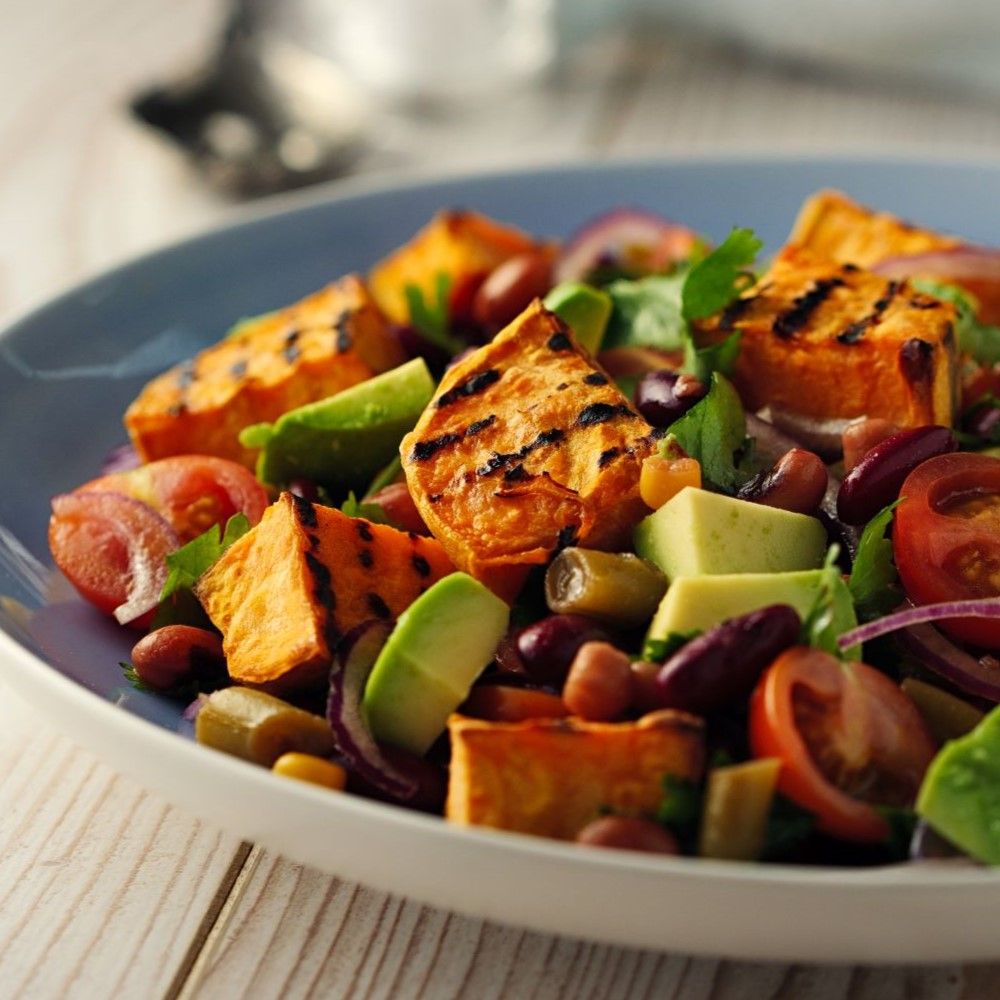 Salat mit gegrillten Süßkartoffelstücken, Avocado, Tomaten, roten Zwiebeln und Bohnen in blauer Schüssel.