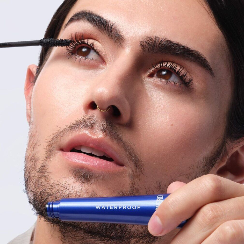 Mann trägt Mascara mit blauer Mascarahülse. Aufschrift: WATERPROOF. Marke: IT Cosmetics.