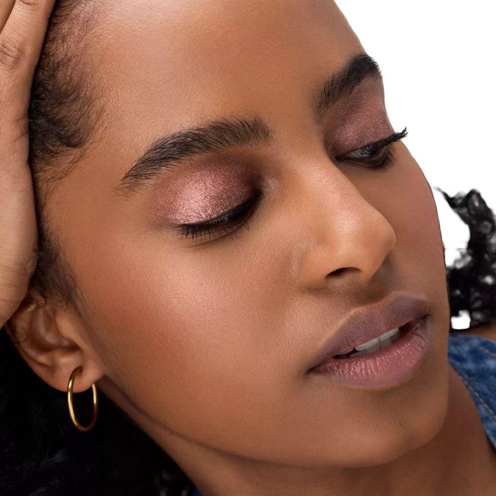 Nahaufnahme einer Frau mit rosafarbenem Lidschatten und goldenen Creolen. Sie stützt den Kopf.