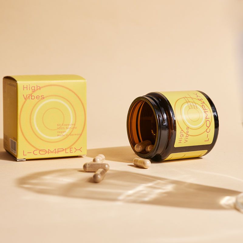 Braune Glasdose und Schachtel mit Kapseln. Dose mit Etikett "High Vibes" und "L-COMPLEX". Kapseln liegen verstreut.